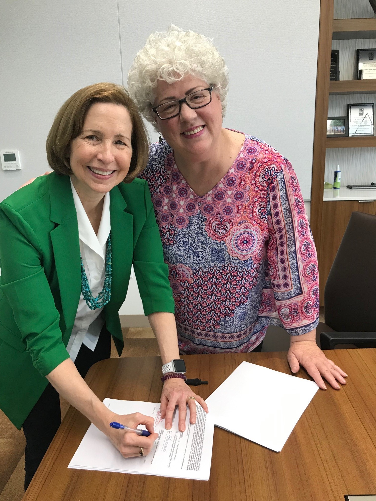 Two women smiling while signing a document in a meeting room.