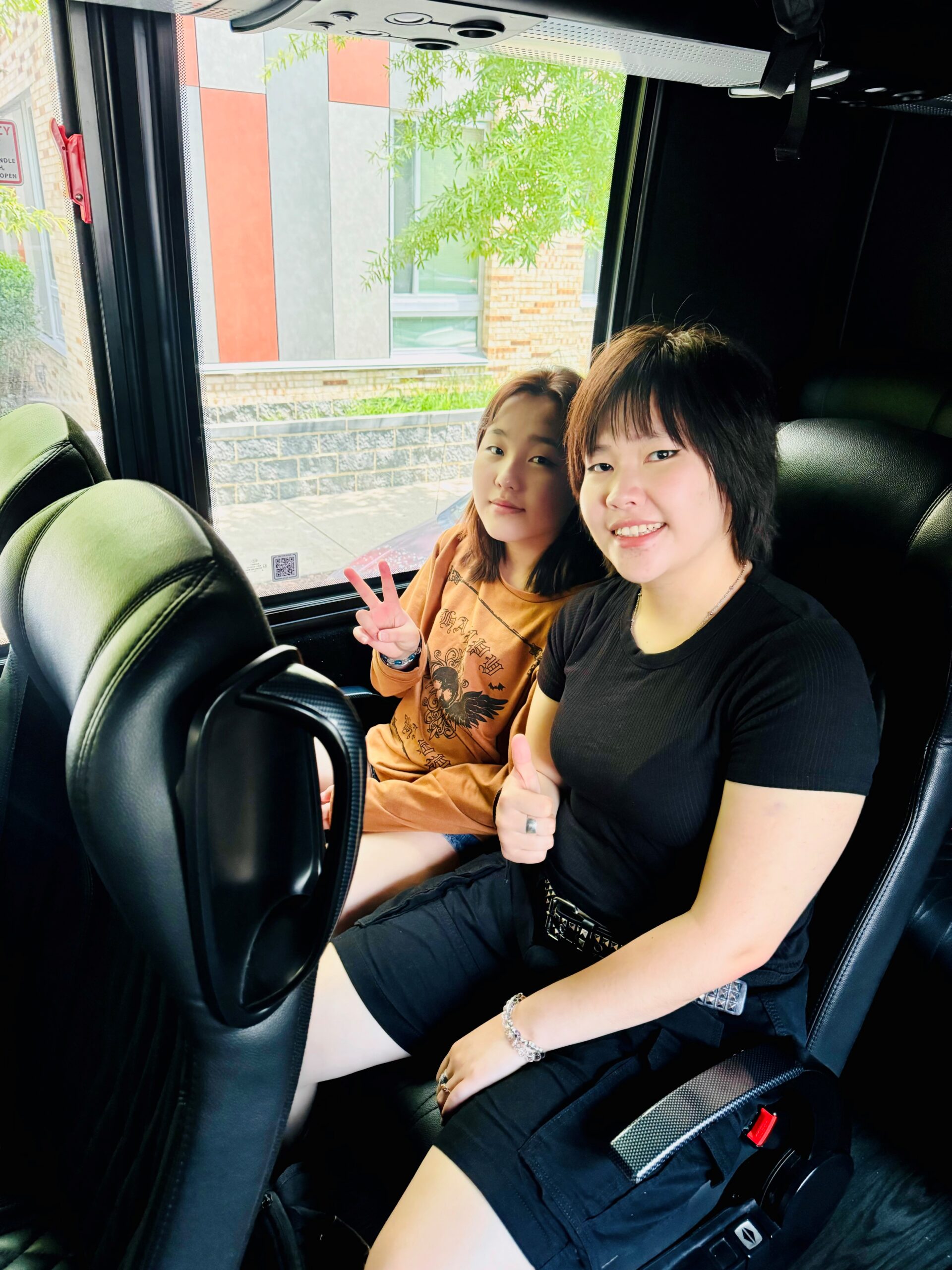 Two girls sitting on a bus with black seats, smiling for the photo.