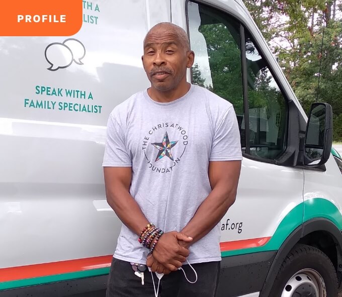 Man in t-shirt and wristbands stands before a van with trees in the background.