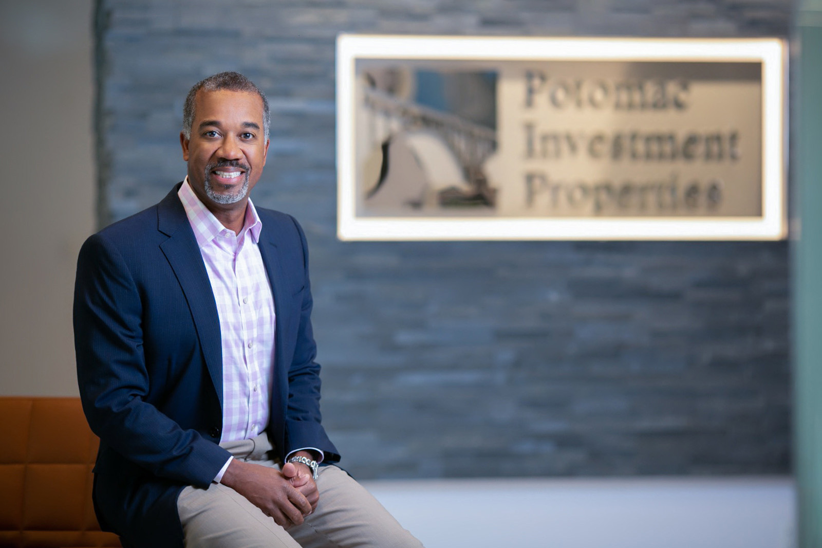 Rich Jordan, smiling for the photo, dressed in a jacket and shirt, sitting on a piece of furniture.