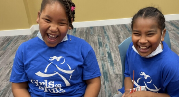 Two young girls, smiling and wearing "Girls on the Run" t-shirts, are sitting together indoors, enjoying their time.