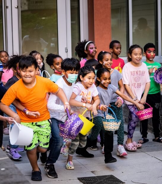 A group of children, holding colorful baskets, excitedly participate in an outdoor event, likely an egg hunt or similar activity.