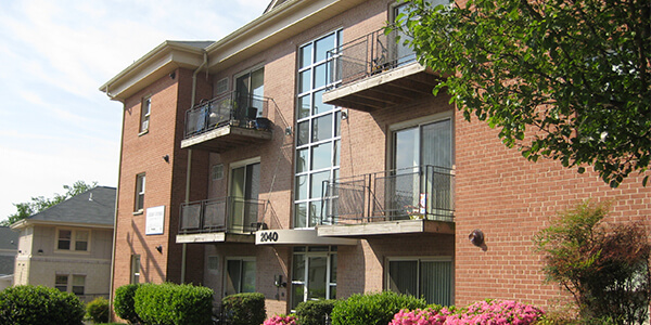 A three-story brick apartment with balconies, large windows, surrounded by greenery and pink flowers.