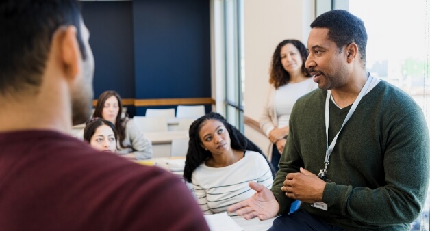 A group of people in a classroom setting, with one man speaking while others listen attentively.