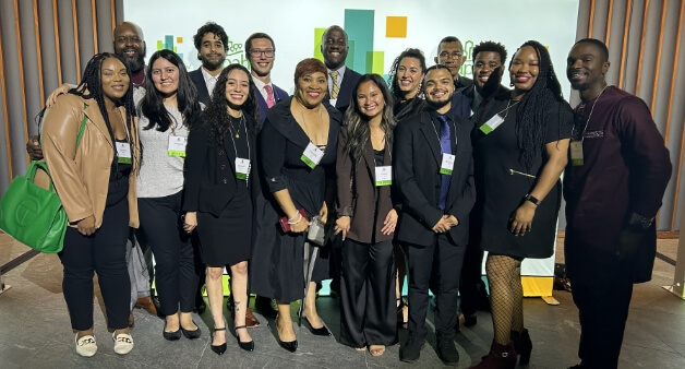 A diverse group of people, dressed in business attire, pose together and smile for a group photo at an event, with conference badges visible.