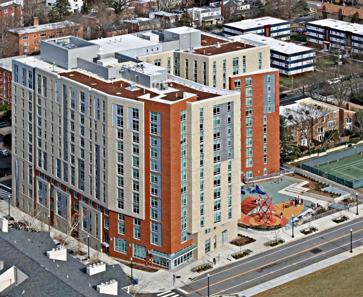 Aerial view of a modern multi-story residential building with a playground and adjacent street.