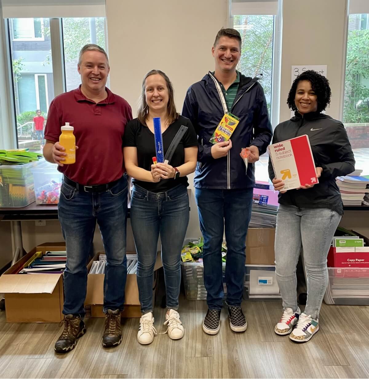 Two men and two women standing, holding school supplies in their hands.