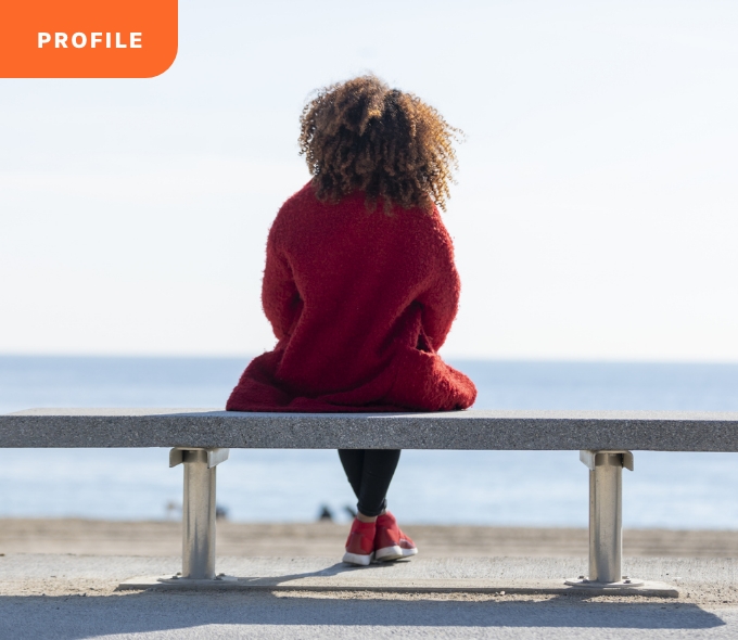 Curly-haired person in red sweater on bench, facing ocean, "Profile" tag top left.