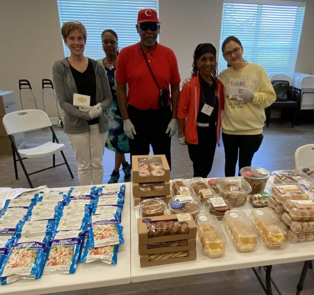 Five people stand behind a table of packaged food in a room with chairs and windows.