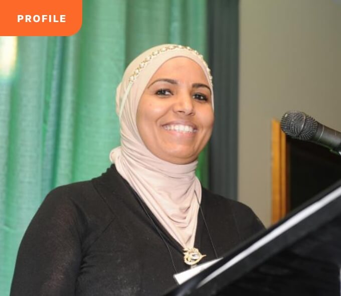 A smiling person in a hijab speaks at a podium with a mic; "PROFILE" label in the top-left corner.