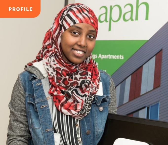 A smiling person in a headscarf and denim jacket stands before a grand building; banner reads "Who We Serve.